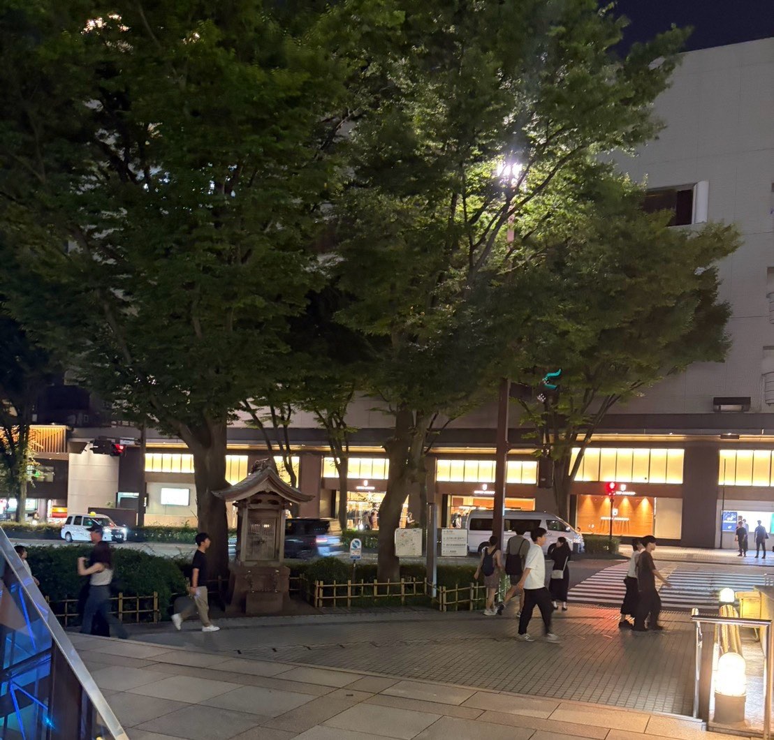 夜の香林坊。街路樹の下に小さな祠があり、奥に明かりのついた商業ビル、右手前に地下道の入口